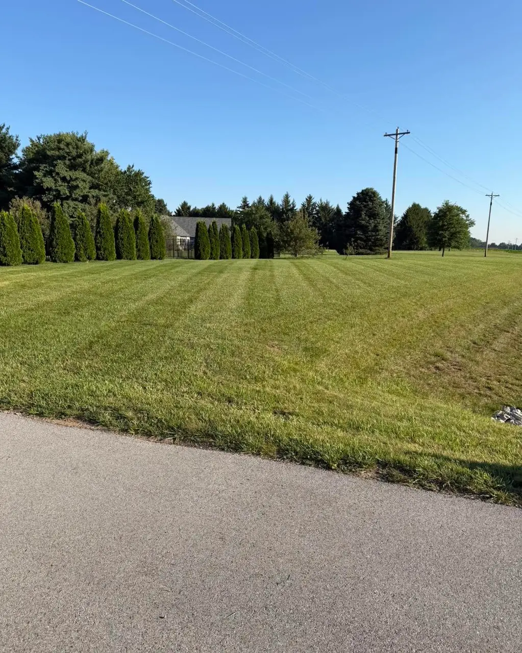 A large open rural lawn mown in long stripes beside a country road, with a power line and tree line in the distance