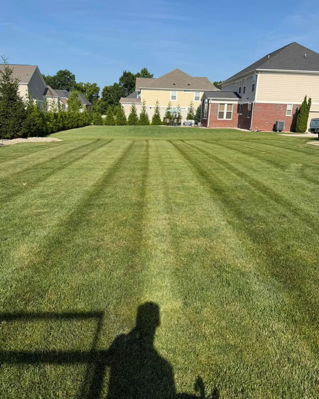 An open striped lawn between houses in a newer subdivision, the photographer's shadow in the foreground