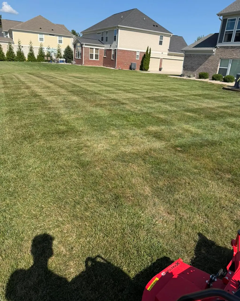 A broad striped lawn running toward a row of houses, with the mower and operator's shadow across the foreground